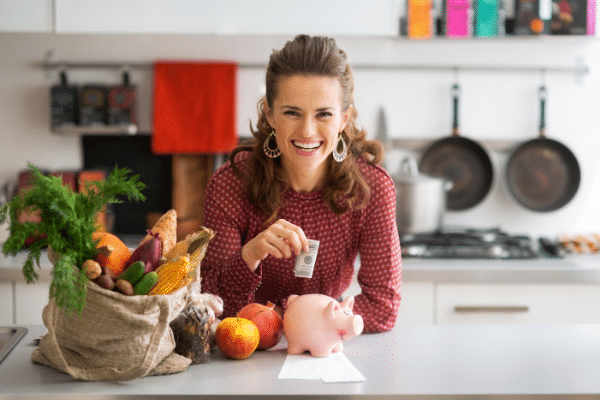 woman in kitchen with groceries putting money in a piggy bank