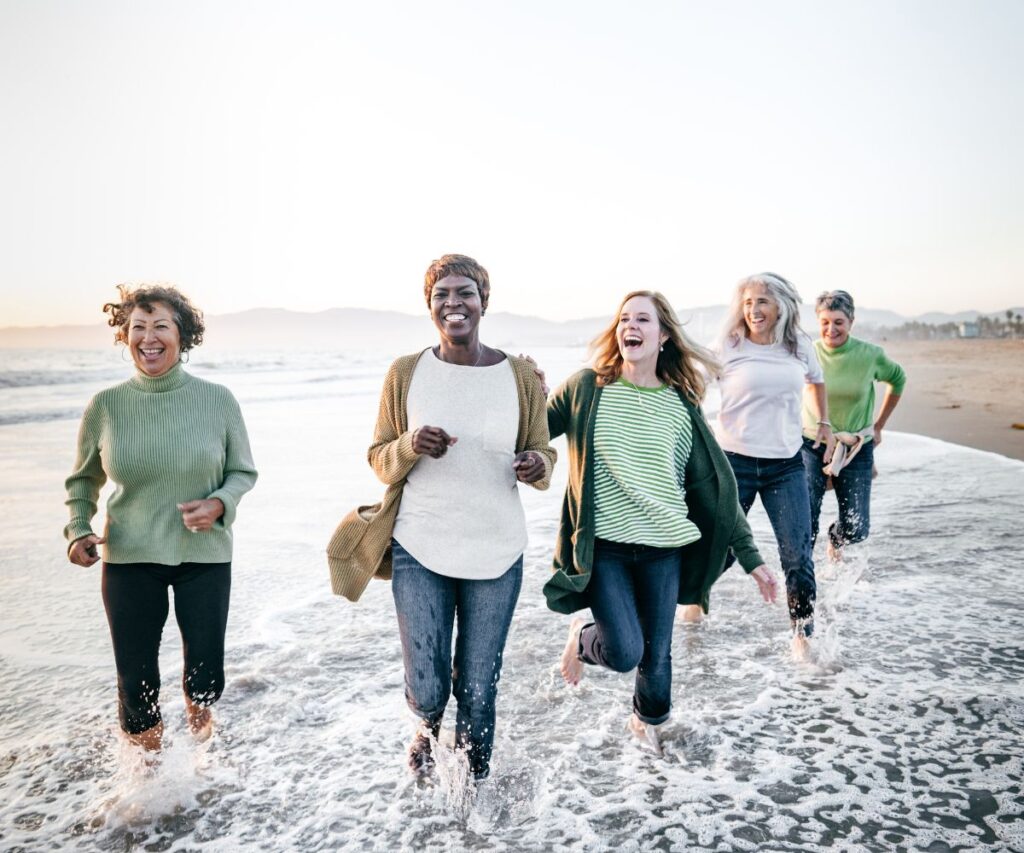 group of five laughing older women wearing sweaters and jeans jogging through the surf on the beach